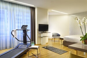 A hotel room with a treadmill, a flat-screen TV, a bed, a chair, and a potted orchid.