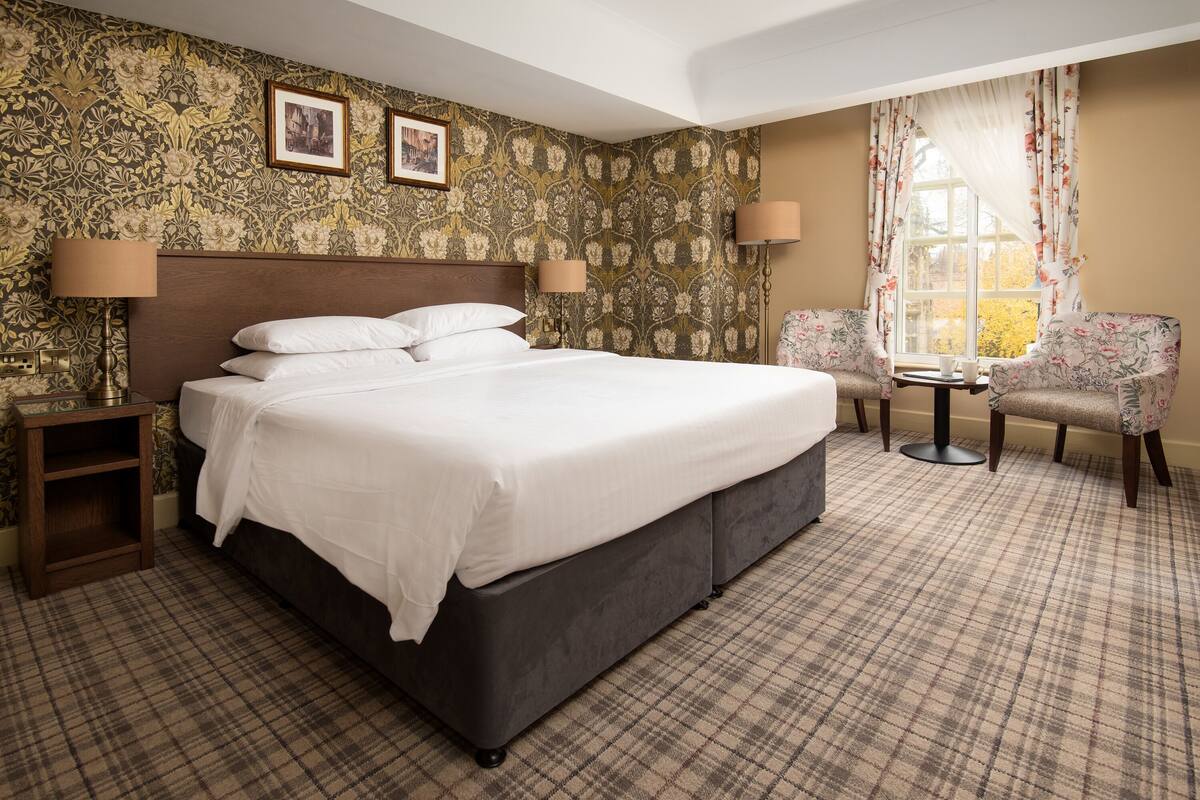 Une chambre d’hôtel avec un grand lit, une chaise, une petite table et une fenêtre avec des rideaux.
