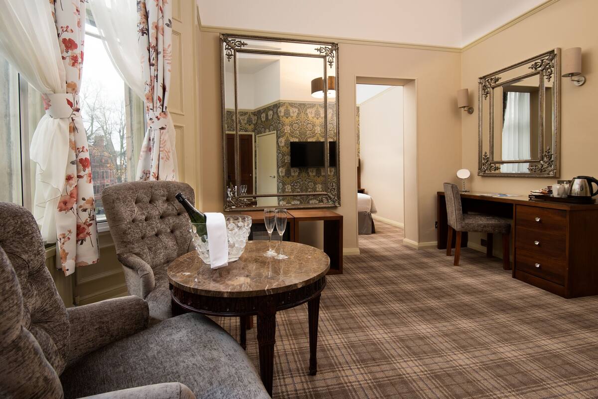 Une chambre d’hôtel avec un grand miroir, une table ronde avec deux verres à vin, un canapé, un bureau avec une chaise et une commode.