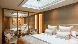 A modern hotel room with a large bed, a sitting area with a chair, a small table with a vase of flowers, and a skylight above the bed.
