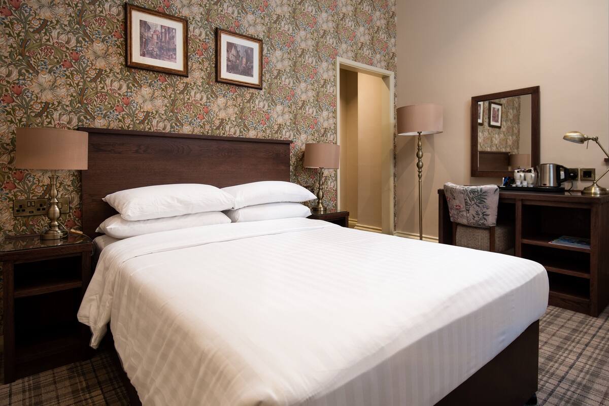 Un lit bien fait, avec du linge de lit blanc, une tête de lit en bois et deux lampes de chevet. On y trouve également une chaise avec un coussin à motifs floraux, un bureau avec une lampe et une machine à café.