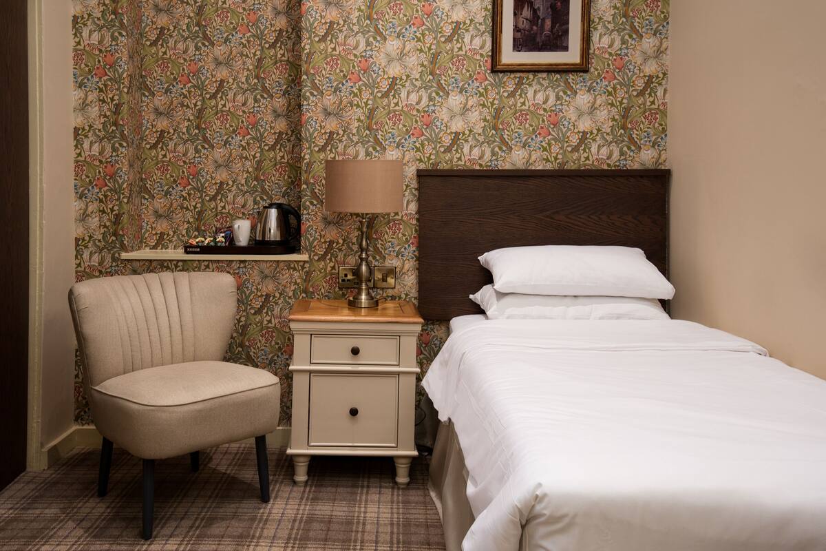 Une chambre d’hôtel avec un lit, une chaise, une table de chevet avec une lampe et une étagère avec une cafetière.