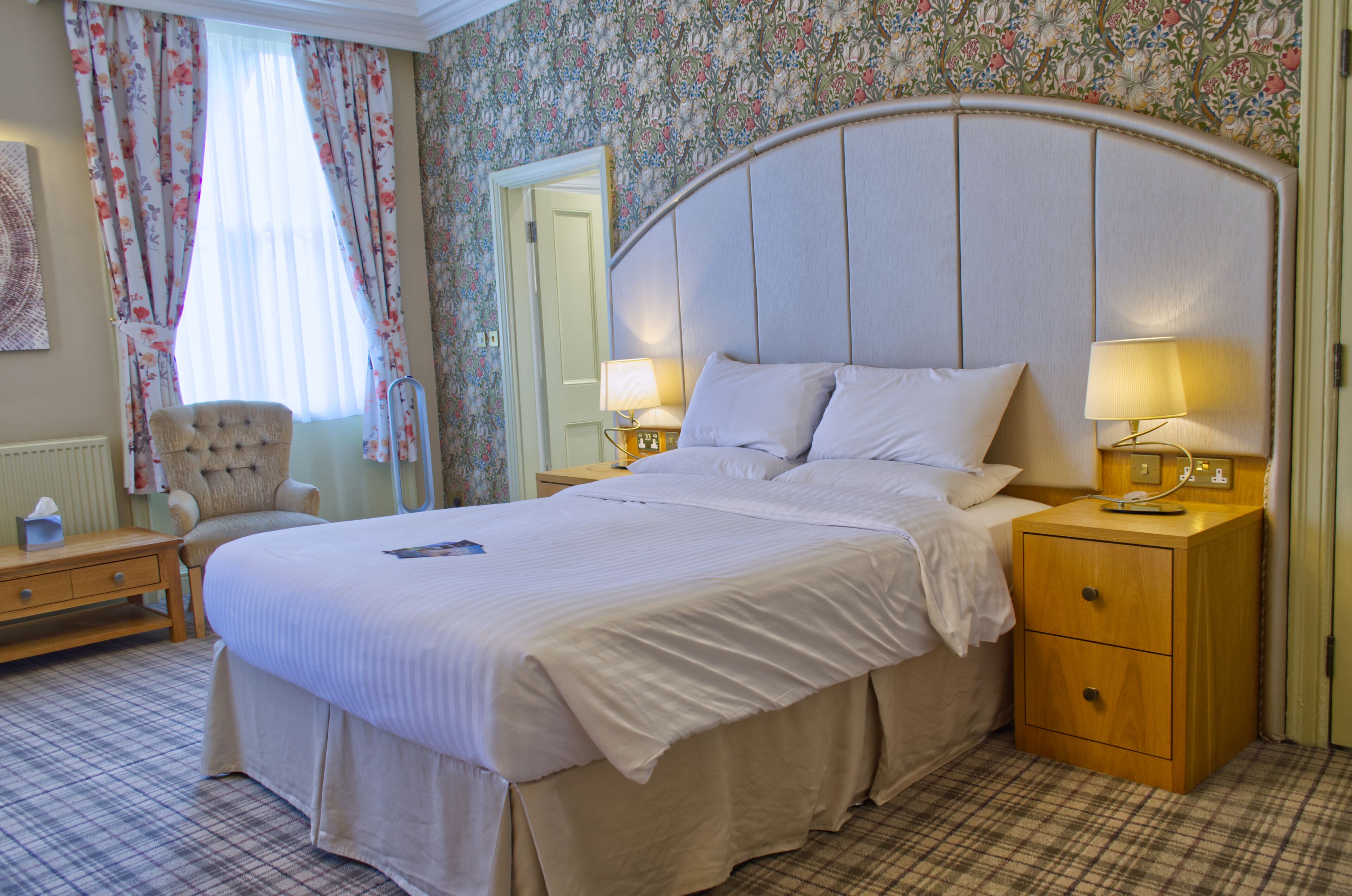 Een net opgemaakt bed met witte lakens, een houten nachtkastje met een lamp, een stoel en een wand met bloemenbehang.