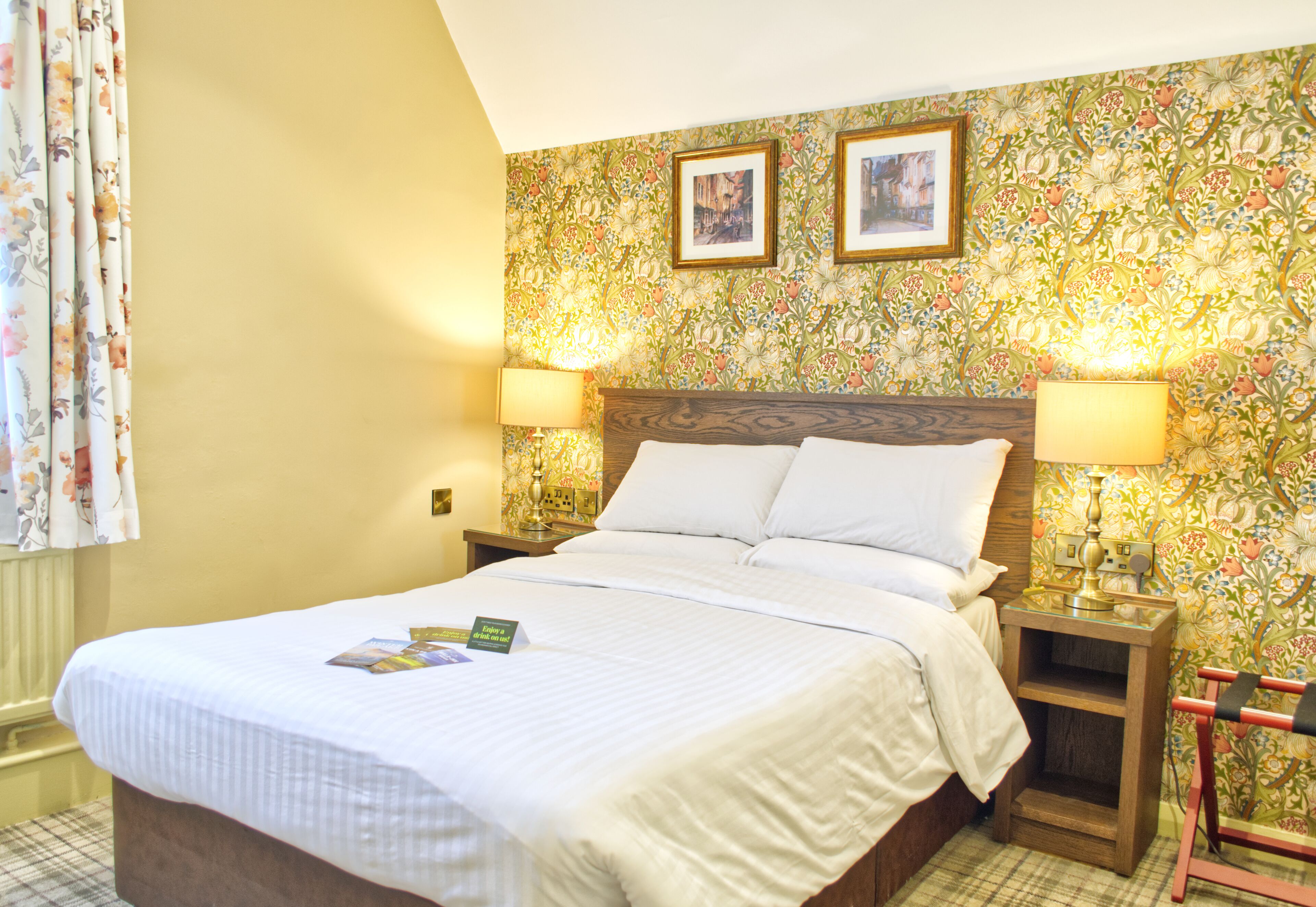 Cama bem arrumada com edredom branco e travesseiros, com duas luminárias de cabeceira e uma pequena mesa de cabeceira de madeira.