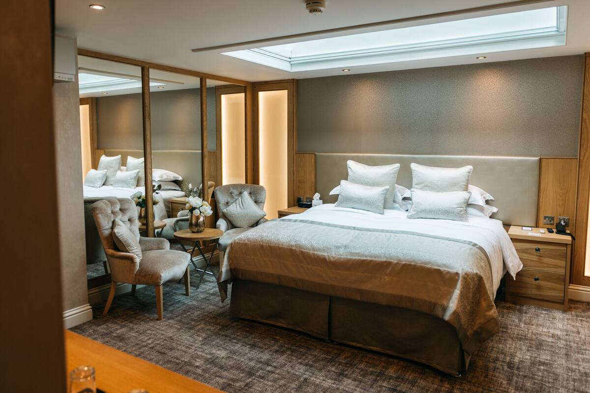 Une chambre d’hôtel moderne avec un grand lit, un coin salon comprenant un fauteuil et une petite table, une table de chevet et un grand miroir.