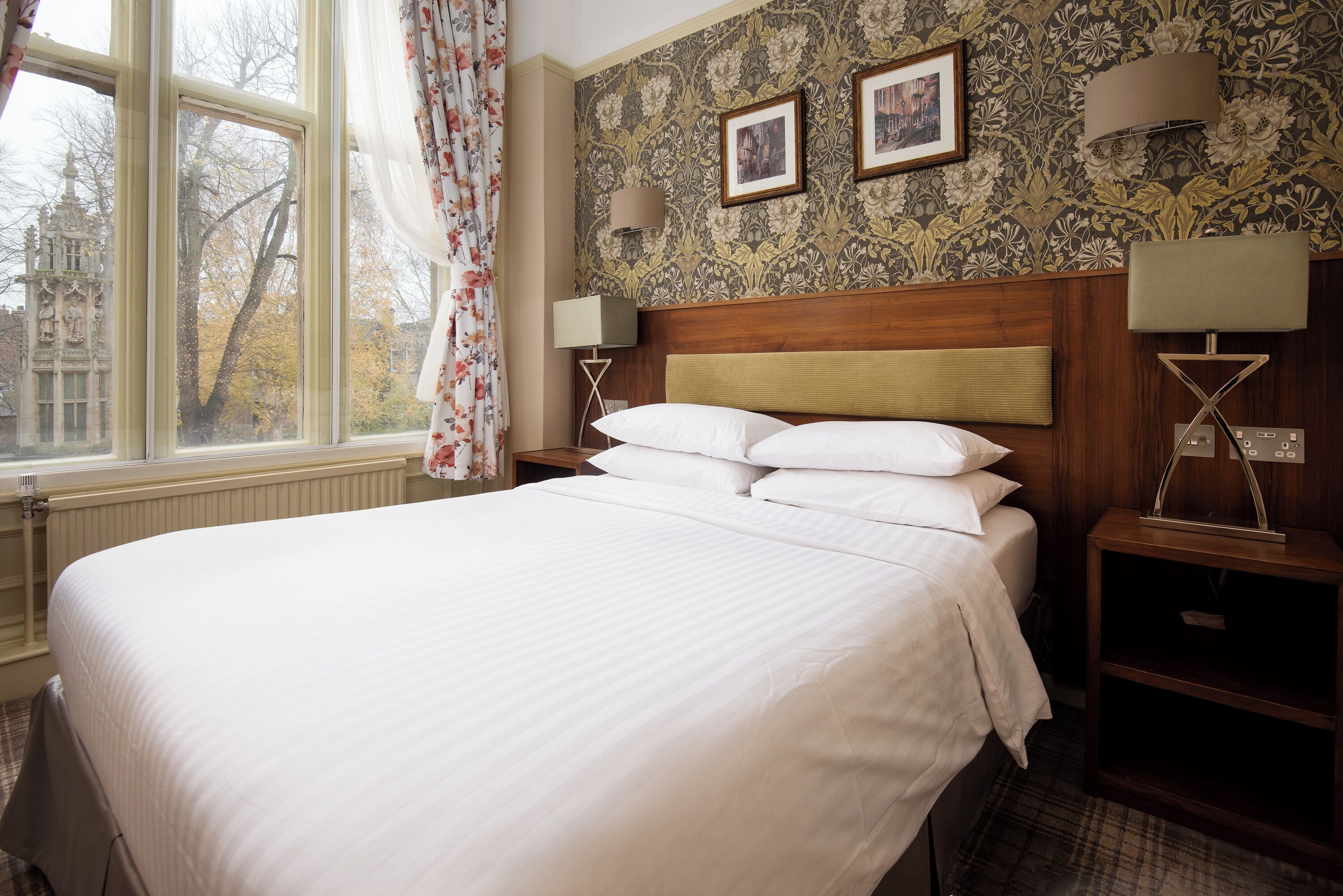 A neatly made bed with white linens, a wooden headboard, and two bedside lamps. A window with floral curtains and a view of trees is visible.