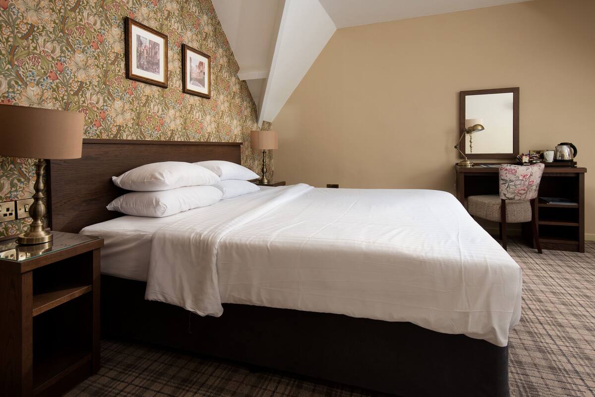 Un lit bien fait dans une chambre d’hôtel, avec un papier peint à motifs floraux, une table de chevet en bois avec une lampe, une petite table d’appoint et une chaise à motifs floraux.