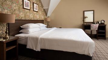 A neatly made bed in a hotel room with a floral wallpaper, a wooden nightstand with a lamp, a bedside table, and a chair with a floral pattern.