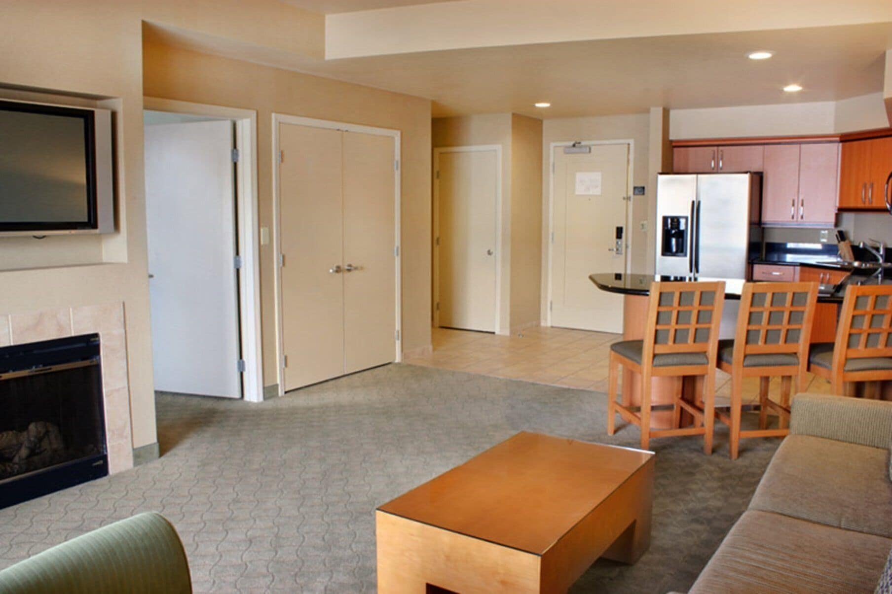 A living room with a fireplace, a dining area with wooden chairs, a kitchen with a refrigerator, and a TV mounted on the wall.