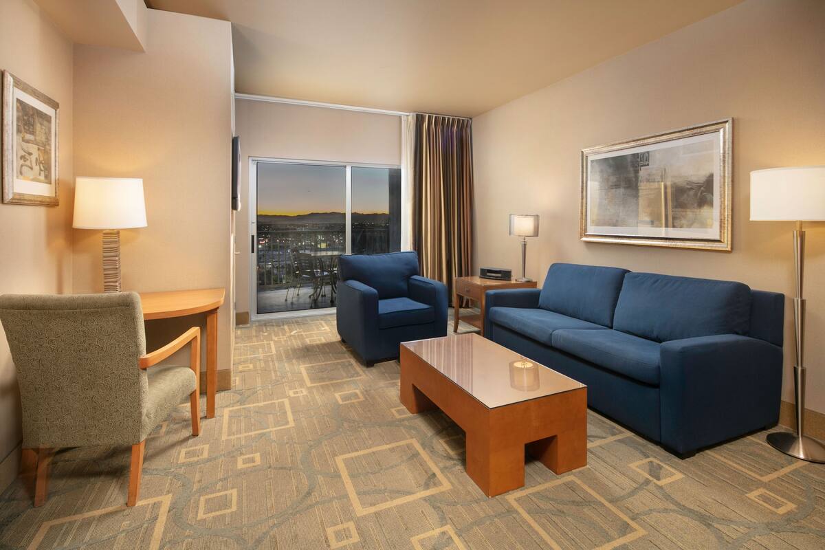 A hotel room with a blue sofa, a wooden coffee table, a patterned armchair, a small table with a lamp, and a large window offering a city view.