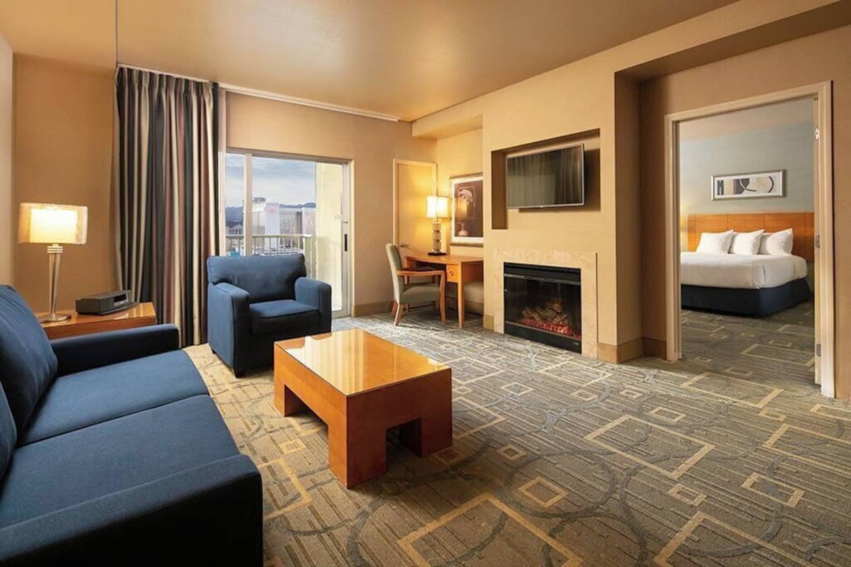 A hotel room with a sofa, armchair, and a small table. There is a fireplace, a desk, and a bed.