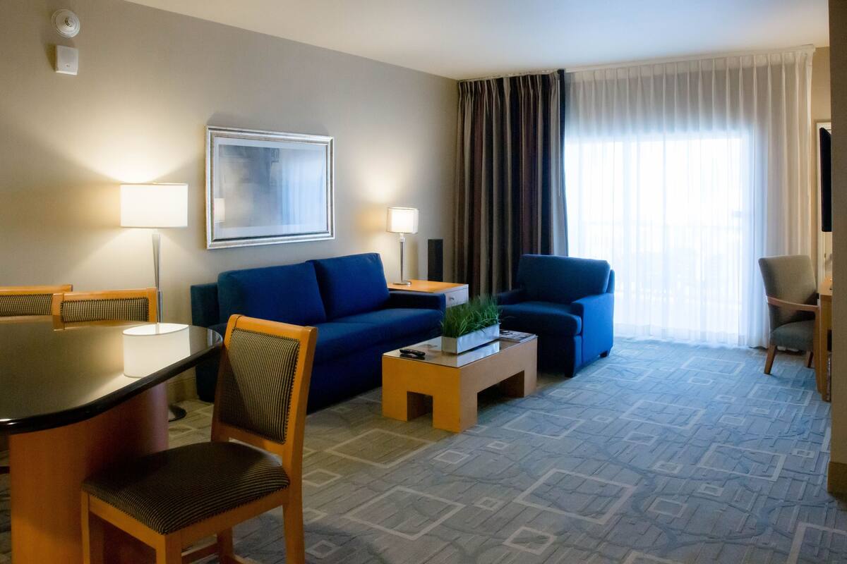 A hotel room with a blue sofa, a small coffee table, a dining table with chairs, and a large window with curtains.