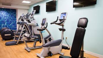 A gym with various exercise equipment, including treadmills and stationary bikes, against a light blue wall with a large TV mounted above.