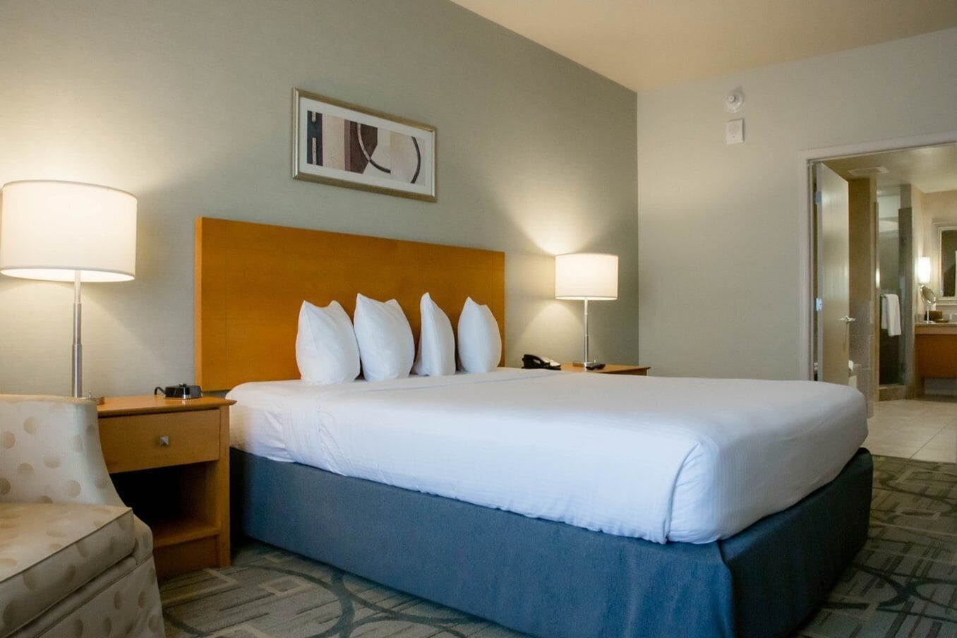 Quarto de hotel com uma cama grande, duas luminárias de cabeceira, mesa de cabeceira, uma cadeira e um quadro na parede.