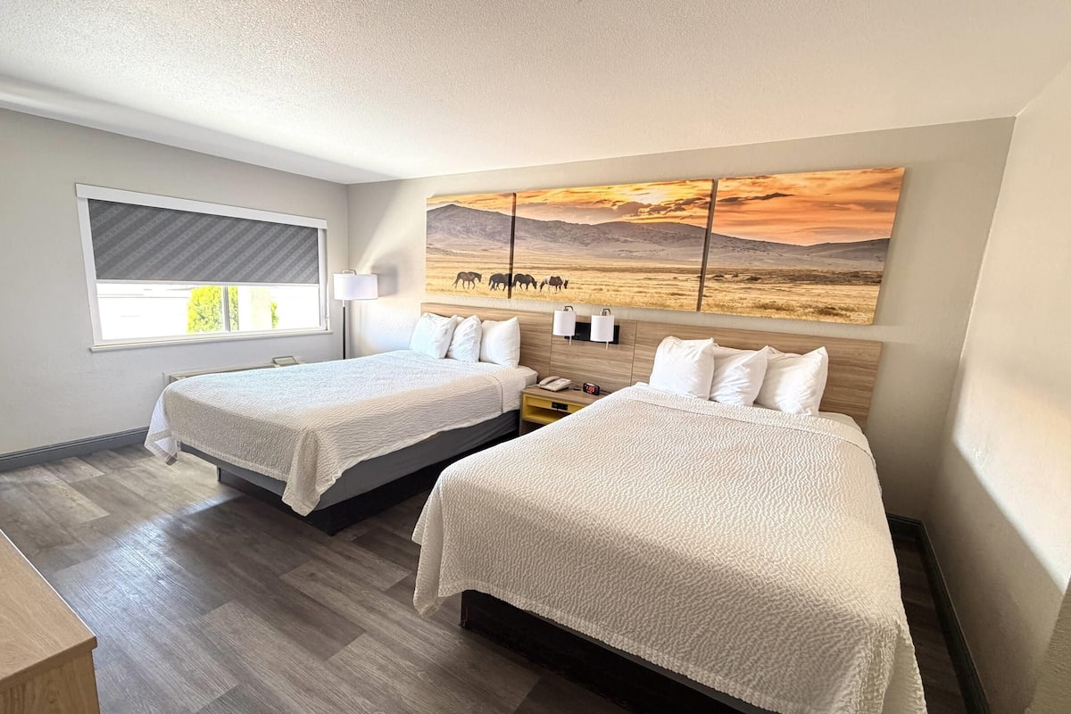 A hotel room with two beds, a large painting of a desert scene, a window with blinds, and a wooden floor.