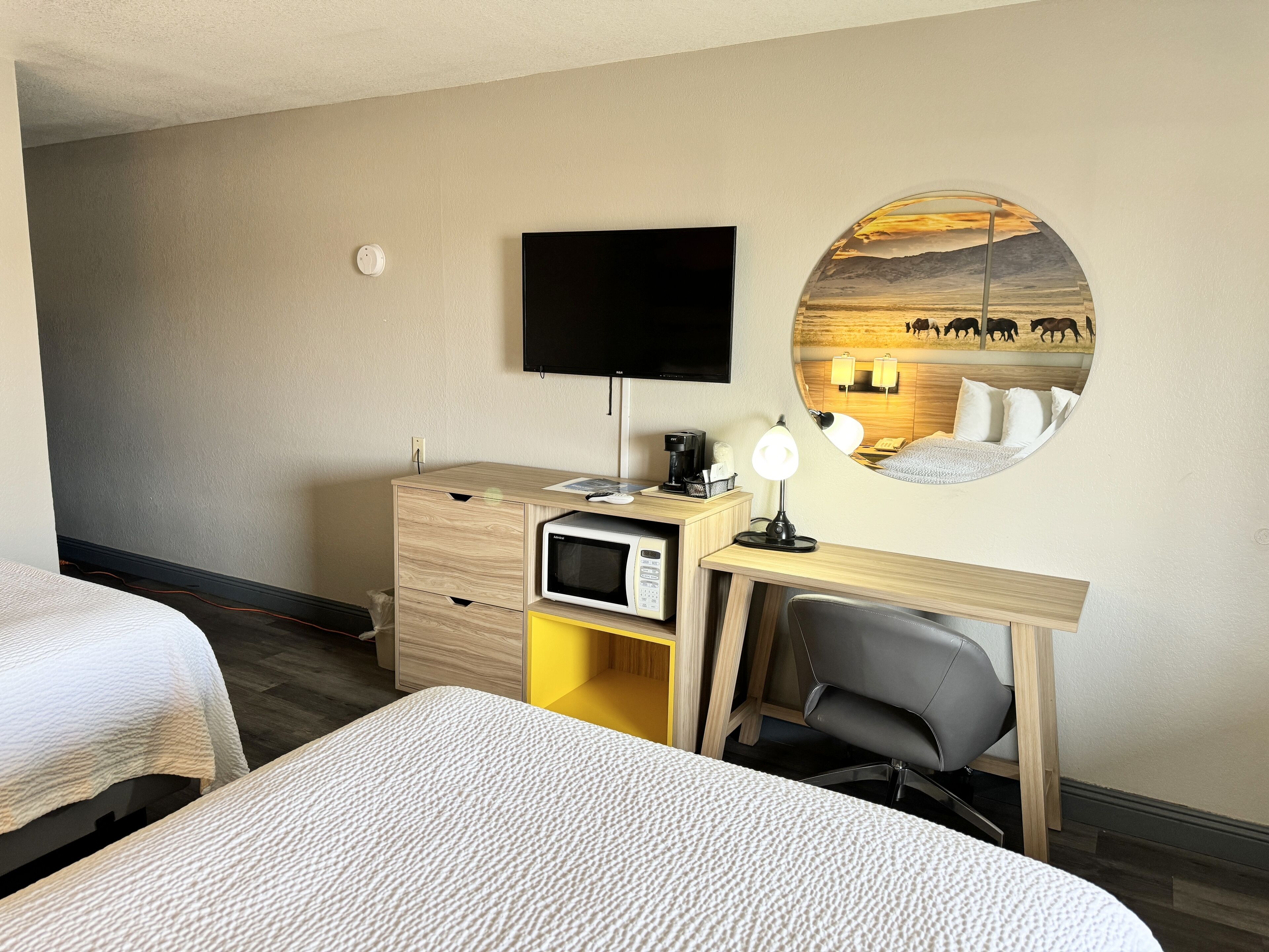 A hotel room with two beds, a desk with a TV, a microwave, a lamp, and a wall-mounted mirror with a scenic painting.