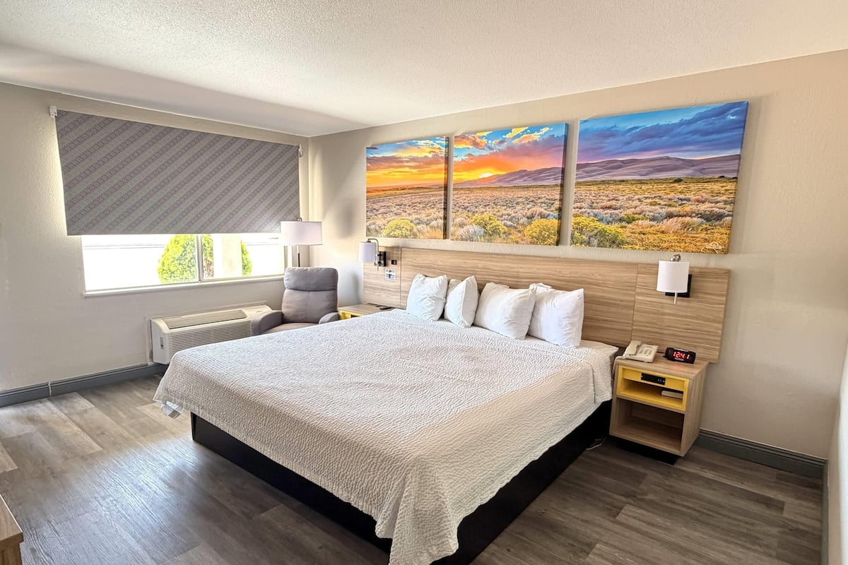A hotel room with a large bed, a nightstand, a lamp, a window with blinds, and two landscape paintings on the wall.
