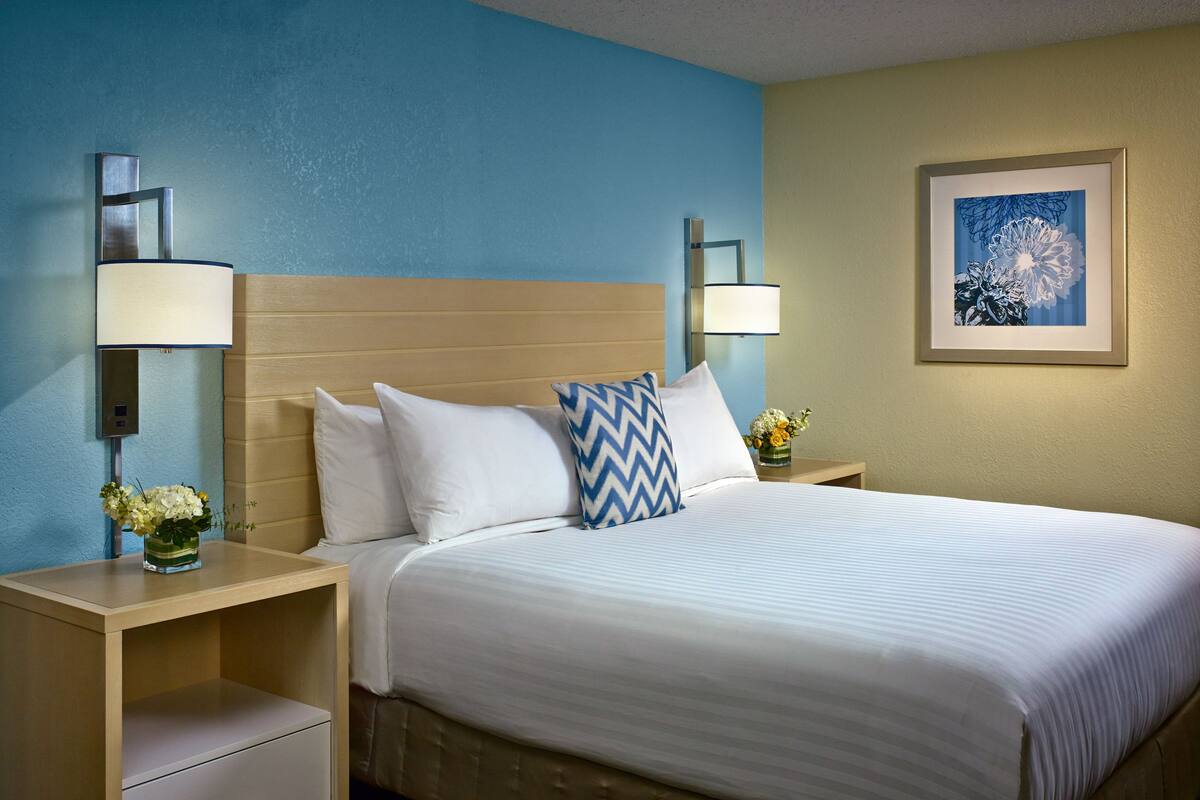 A hotel room with a bed, two nightstands, a drawer, a wall-mounted lamp, a framed picture, and a vase with flowers.