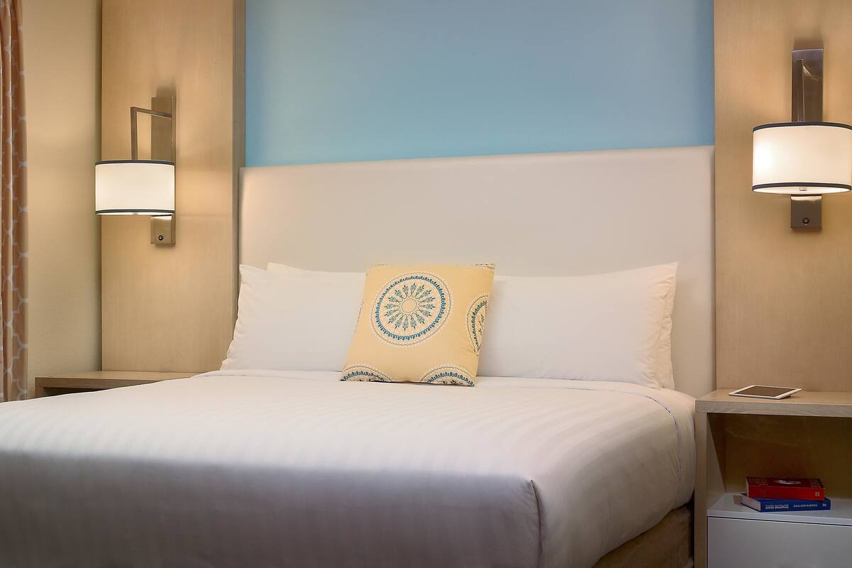 A neatly made bed with white linens, a decorative pillow, and a bedside table with a book and a tablet.