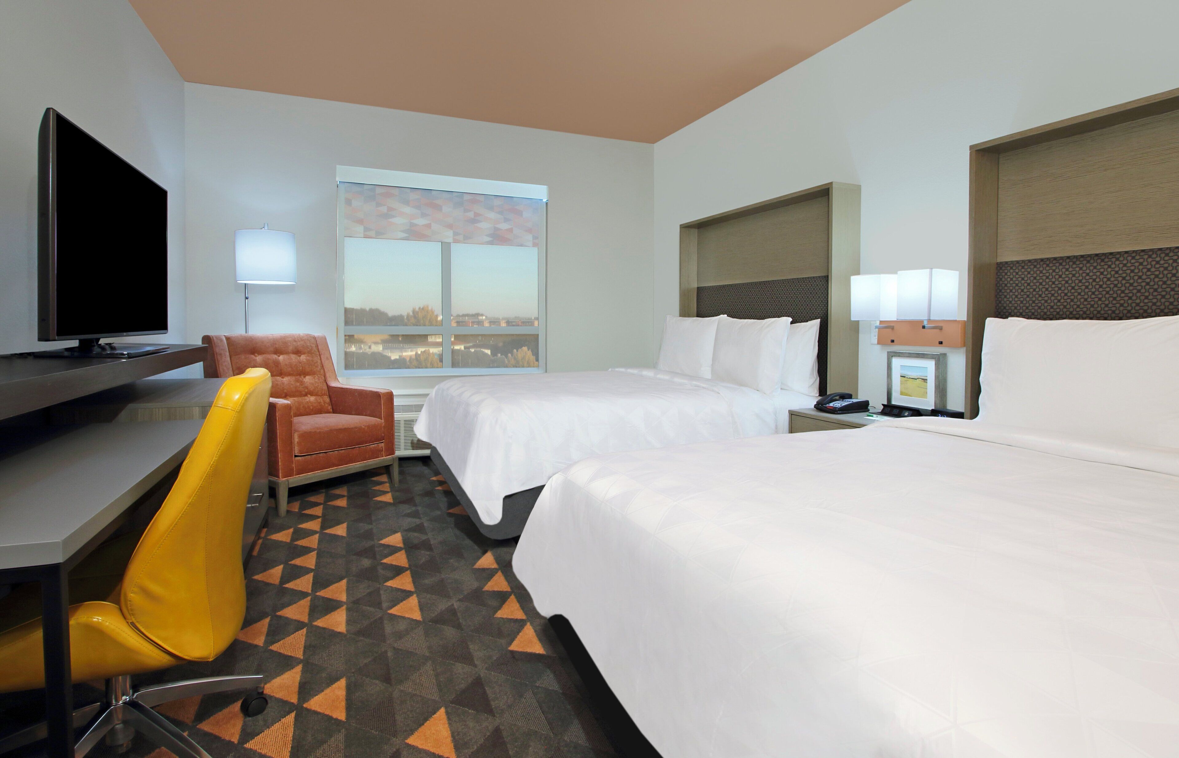 A hotel room with two beds, a desk with a flat-screen TV, a chair, and a window with a city view.