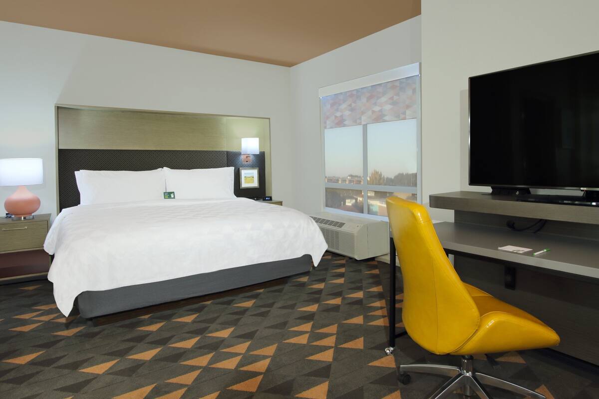 Habitación de hotel con cama, escritorio con televisor de pantalla plana, una silla y ventana con vista a la ciudad.