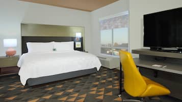 A hotel room with a bed, a desk with a flat-screen TV, a chair, and a window with a city view.