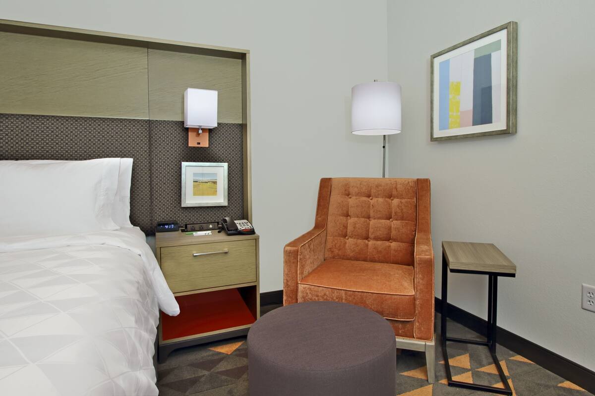 Habitación de hotel con cama, mesita de noche con teléfono, una silla, lámpara y un cuadro en la pared.