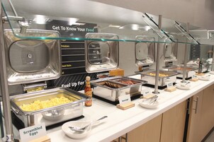 Un bufé con estaciones de comida señalizadas, que incluyen huevos revueltos, salchichas y tocino.
