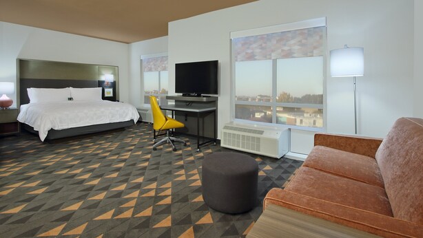 Habitación de hotel con cama, escritorio, televisor, sofá y una ventana grande con vista a la ciudad.