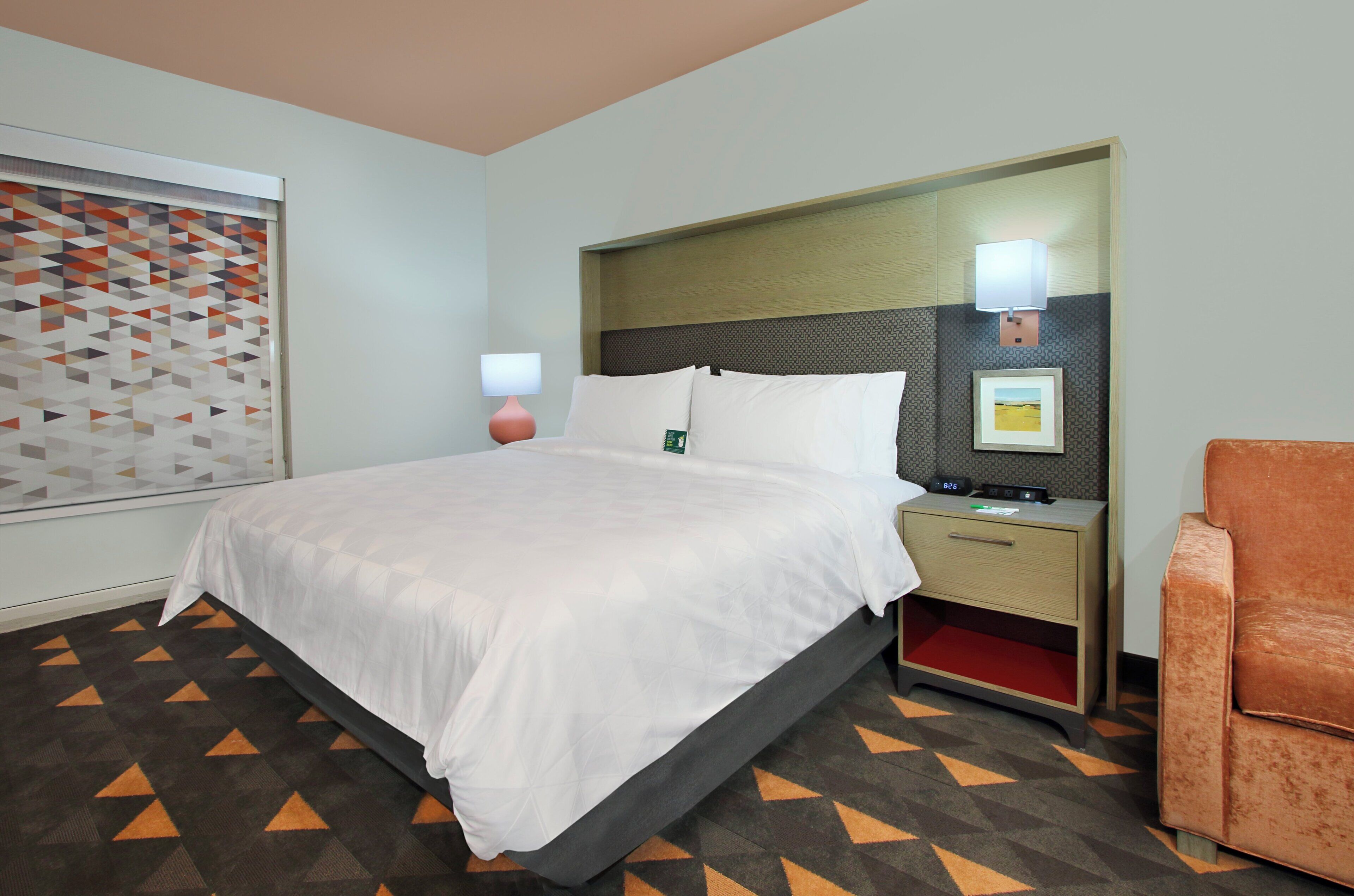 Quarto de hotel com cama, mesa de cabeceira, abajur e uma cadeira.