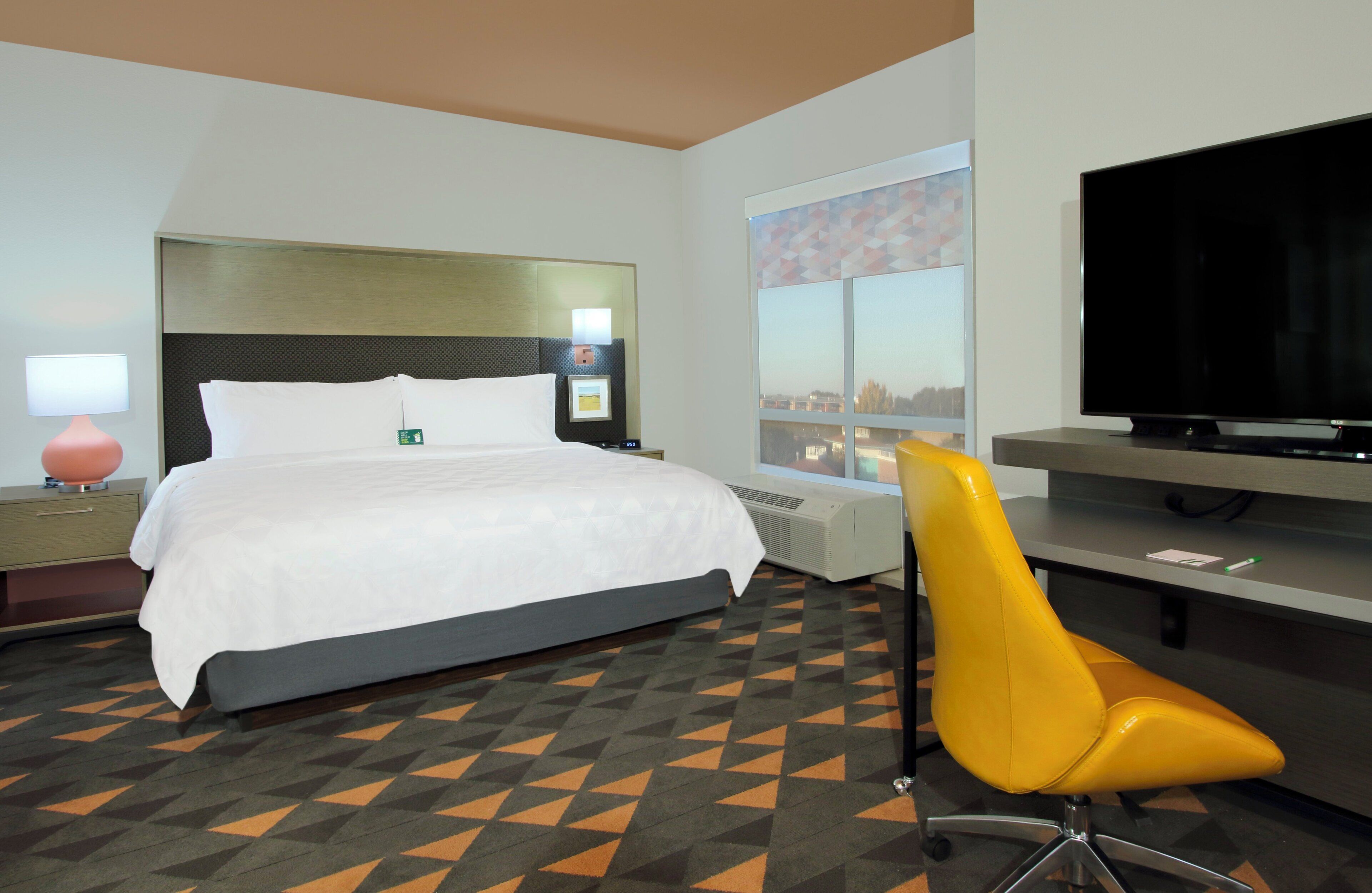A hotel room with a bed, a desk with a flat-screen TV, a chair, and a window with a city view.