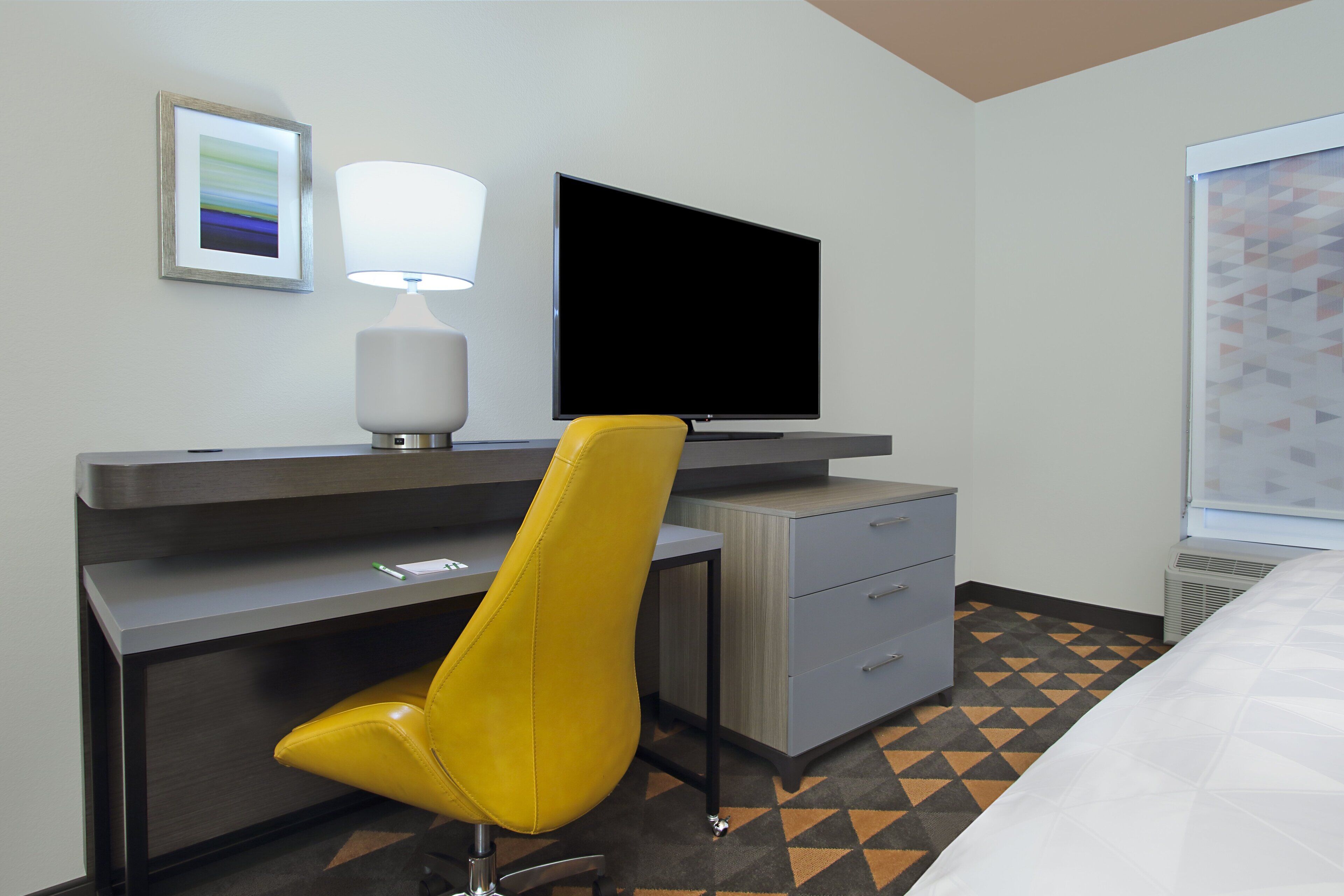 Una habitación de hotel con escritorio, una silla amarilla, televisor de pantalla plana, lámpara, cama y piso con diseño.