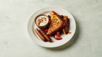 A plate with French toast, sausages, strawberries, blueberries, and a small bowl of yogurt with granola.