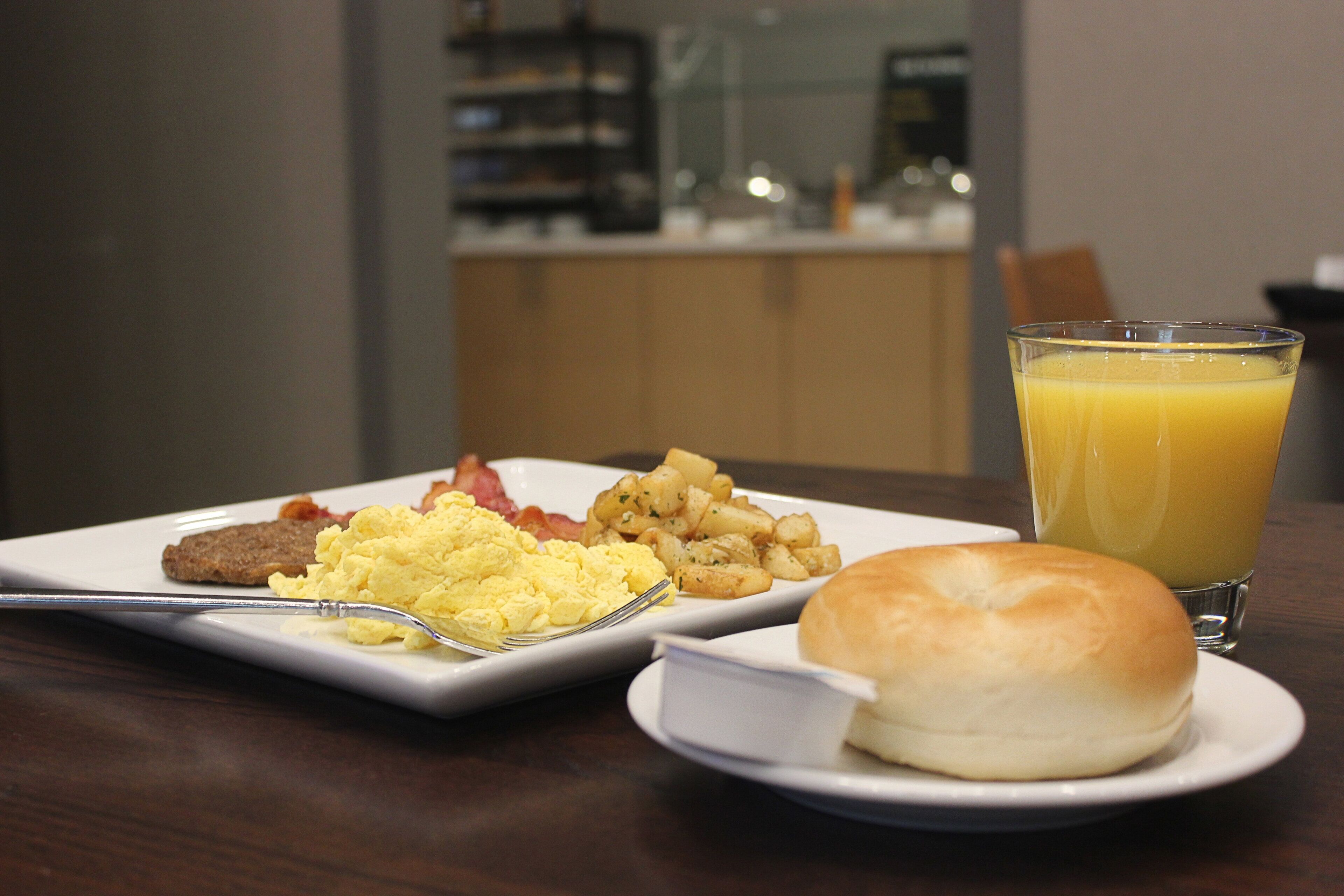 A breakfast plate with scrambled eggs, bacon, sausage, and potatoes, a glass of orange juice, and a roll on a wooden table.