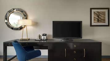 A modern hotel room with a dark wooden desk, a blue chair, a flat-screen TV, a round mirror, a lamp, and a framed picture on the wall.