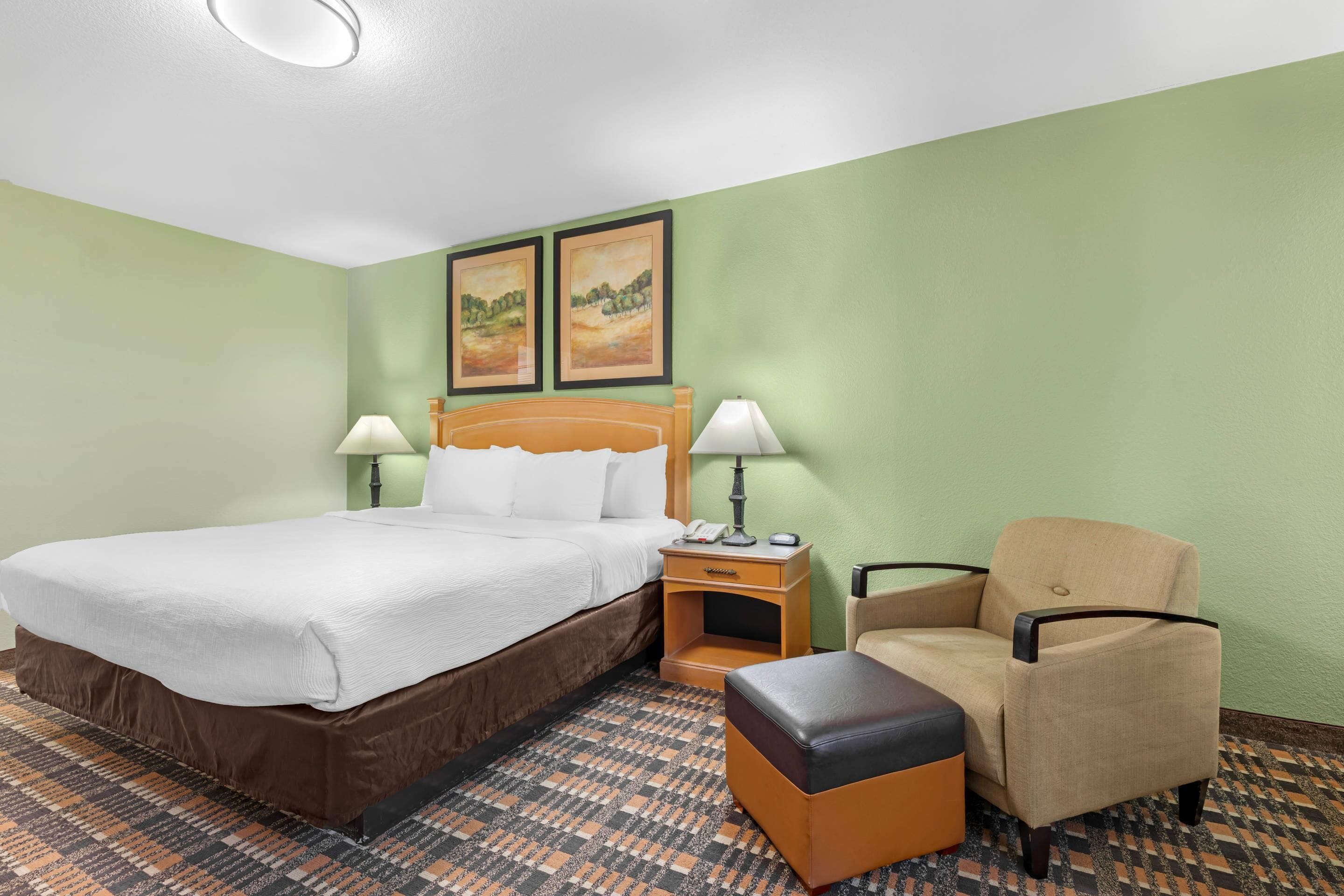 Quarto de hotel com uma cama, duas luminárias de cabeceira, uma cadeira, uma mesinha e duas imagens emolduradas na parede.