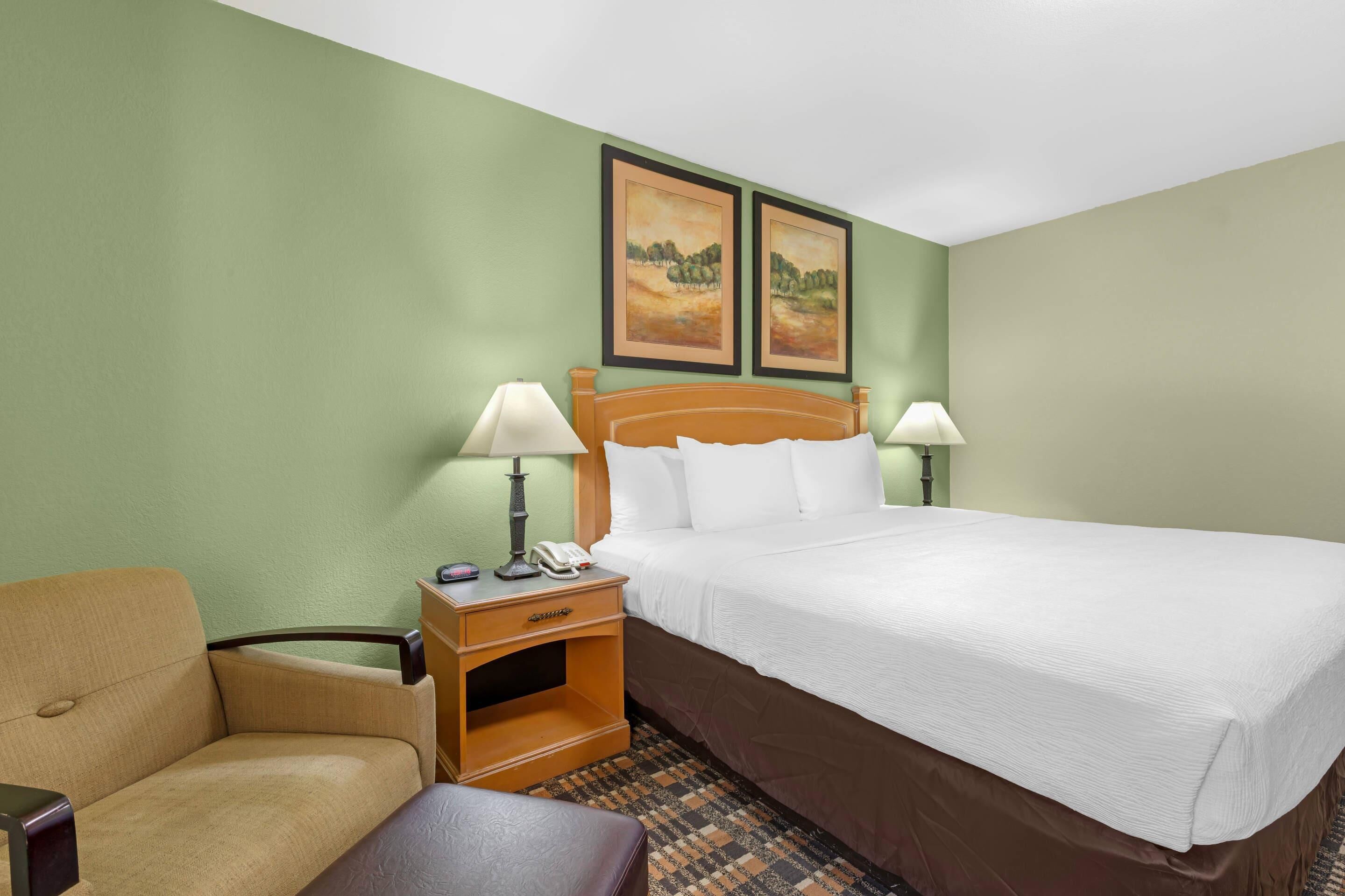 Habitación de hotel con una cama, dos lámparas de noche, una mesita de noche con un teléfono, una silla y dos cuadros de paisajes en la pared.