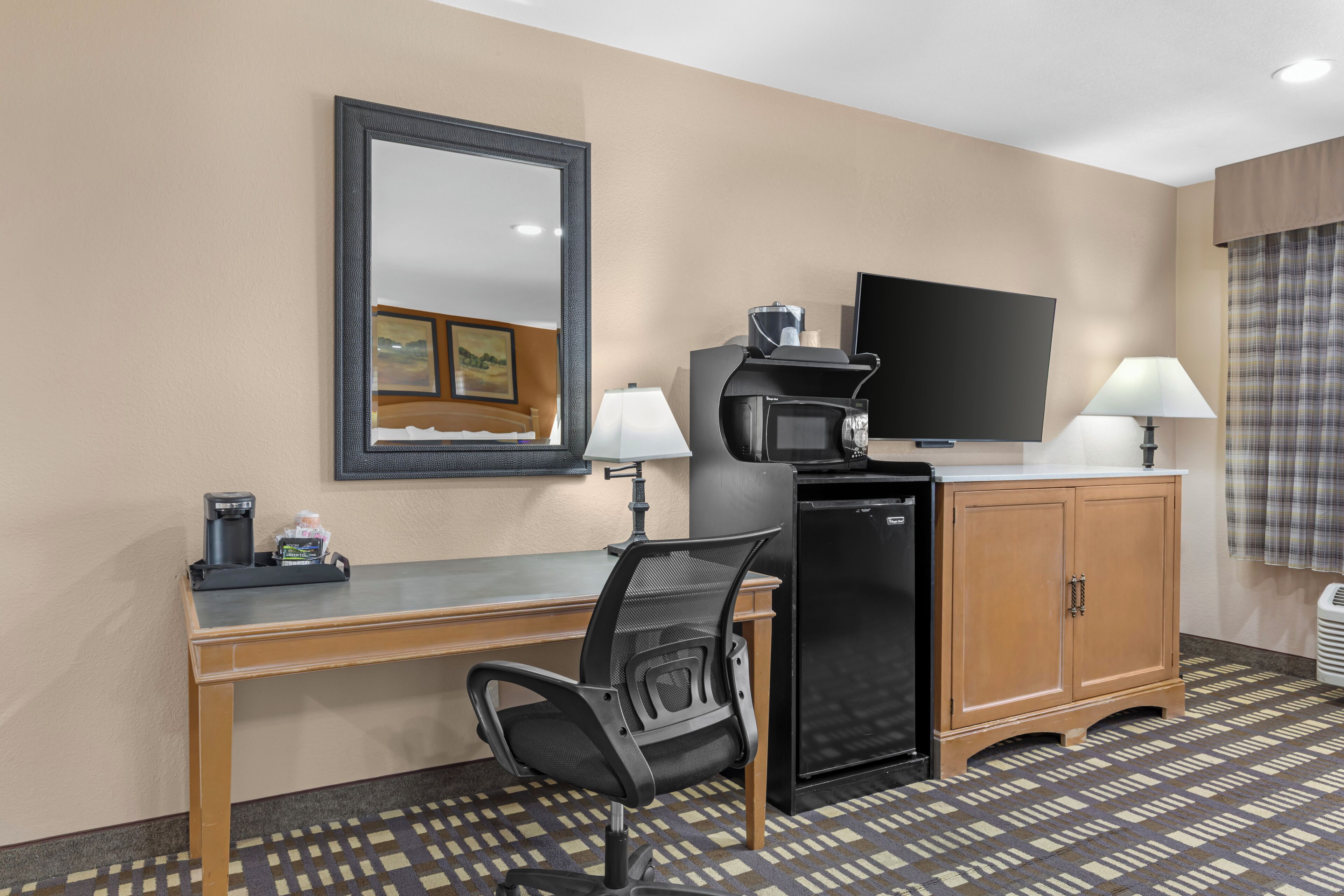 A hotel room with a desk, chair, television, refrigerator, and a patterned floor.