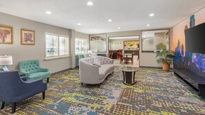 A modern hotel room with a grey sofa, teal armchair, and a glass coffee table. There is a flat-screen TV mounted on the wall, a potted plant, and a patterned carpet. - Quality Inn & Suites Plano East - Richardson (Plano)