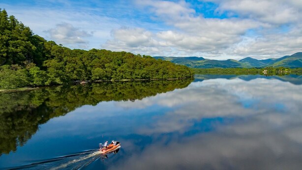 A calm lake with a boat on the water, surrounded by lush greenery and mountains in the background. - Buchanan Arms Hotel & Leisure, Sure Hotel Collection (Drymen)