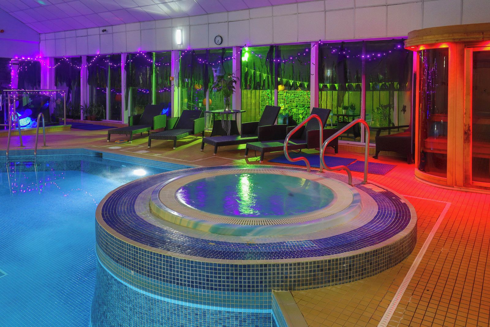An indoor swimming pool with a hot tub, lounge chairs, and a glass enclosure.