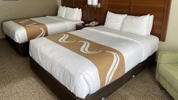 A hotel room with two beds, each with white linens and brown decorative stripes. There is a wooden headboard, a bedside table with a lamp, and a telephone. The room has a carpeted floor and a green chair. - Quality Inn Placentia Anaheim Fullerton (Placentia)