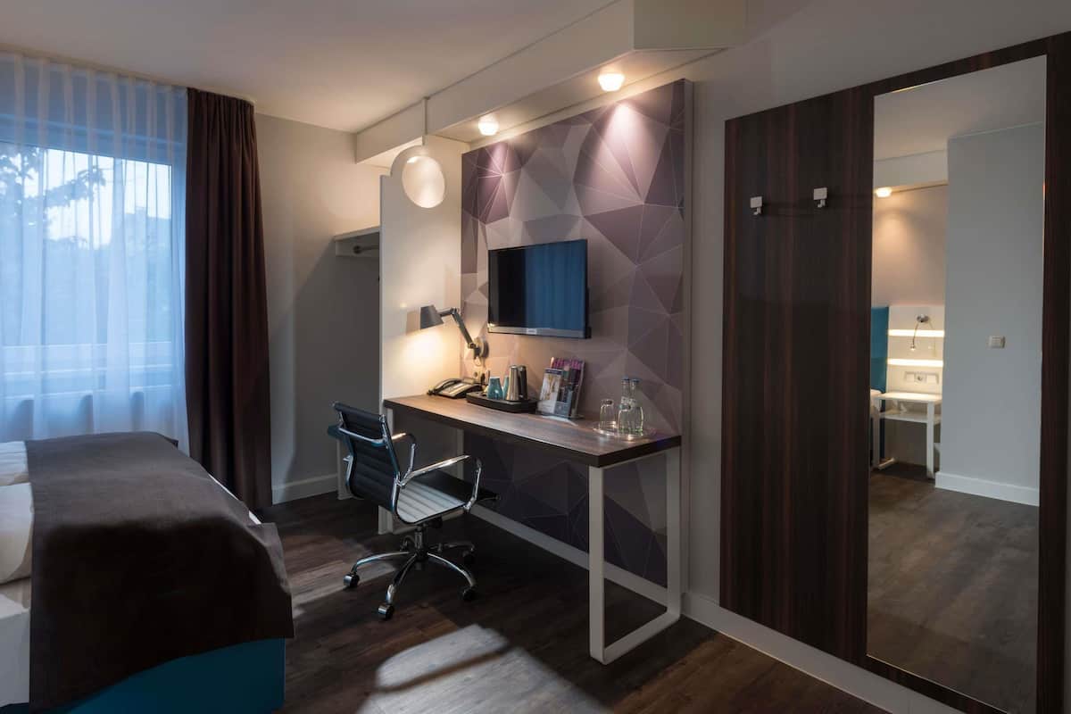 Moderni hotellihuone, jossa on sänky, työpöytä, tuoli, televisio ja peili.