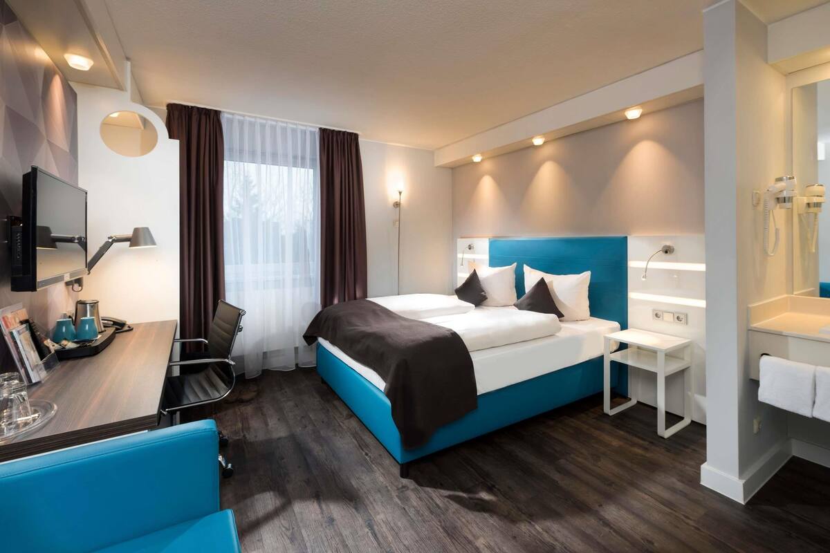 Moderni hotellihuone, jossa on suuri sänky, työpöytä tuolilla, pieni pöytä lampun kanssa, peili sekä kylpyhuone, jossa on pesuallas ja pyyhekoukku.
