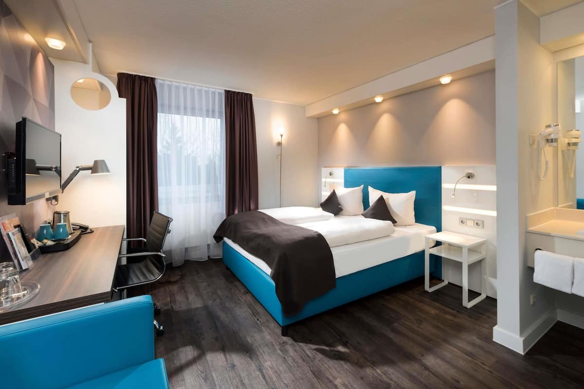 Ein modernes Hotelzimmer mit einem großen Bett, einem Schreibtisch mit Stuhl, einem kleinen Tisch mit Lampe, einem Spiegel und einem Badezimmer mit Waschbecken und Handtuchhalter.