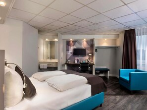 Quarto de hotel com cama, escrivaninha, cadeira, televisão e banheiro.