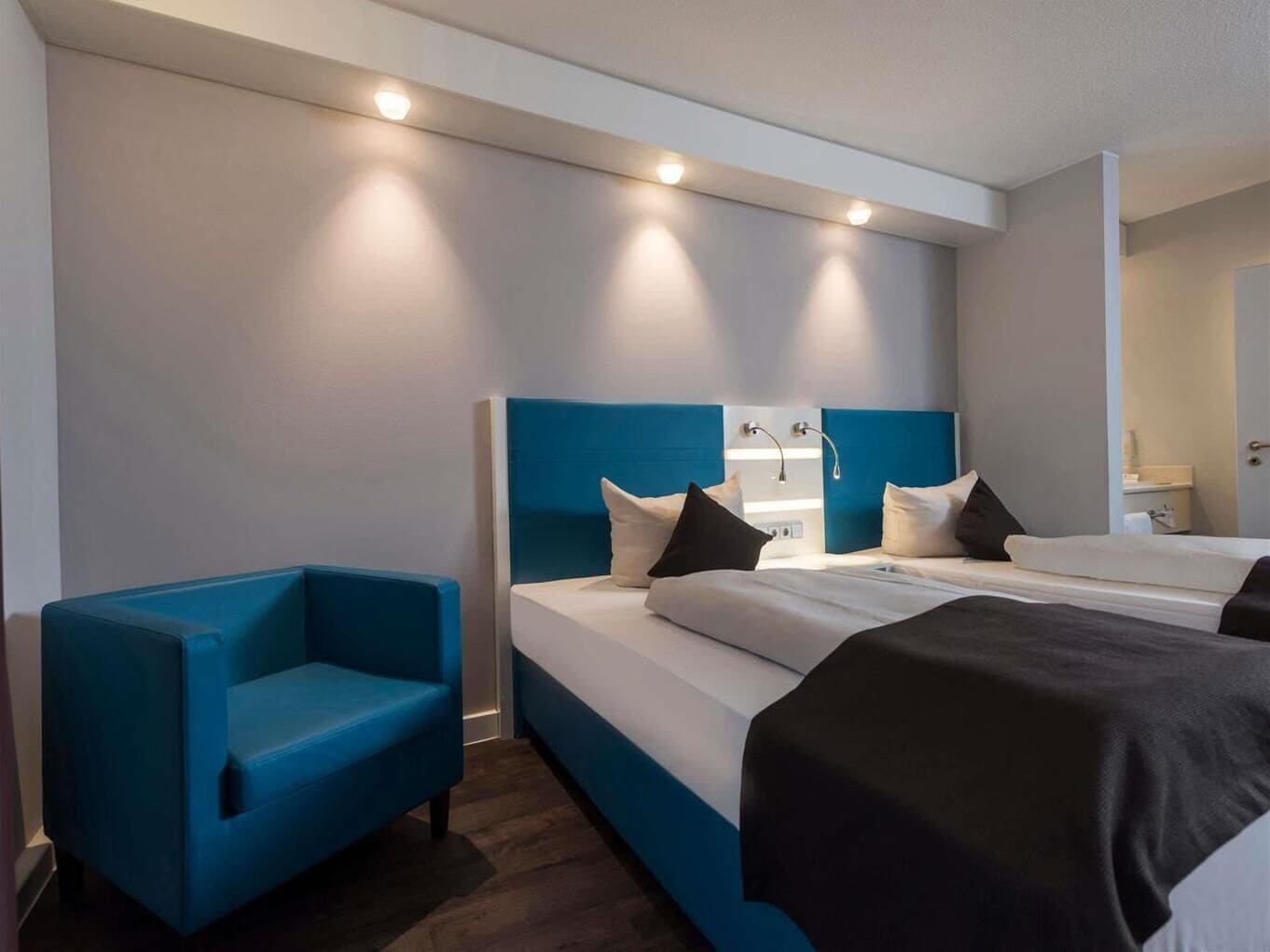 A modern hotel room with a large bed, a blue armchair, and a bedside table.