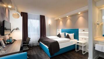 Una habitación de hotel moderna con una cama grande, un escritorio con una silla, una mesita y un baño con espejo y lavamanos.