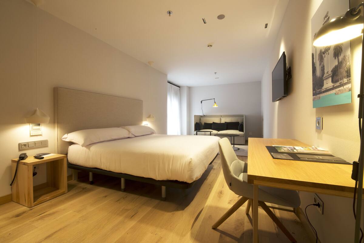 Une chambre d’hôtel moderne dotée d’un grand lit, d’une table de chevet avec une lampe, d’un bureau avec une chaise et d’un téléviseur à écran plat fixé au mur.