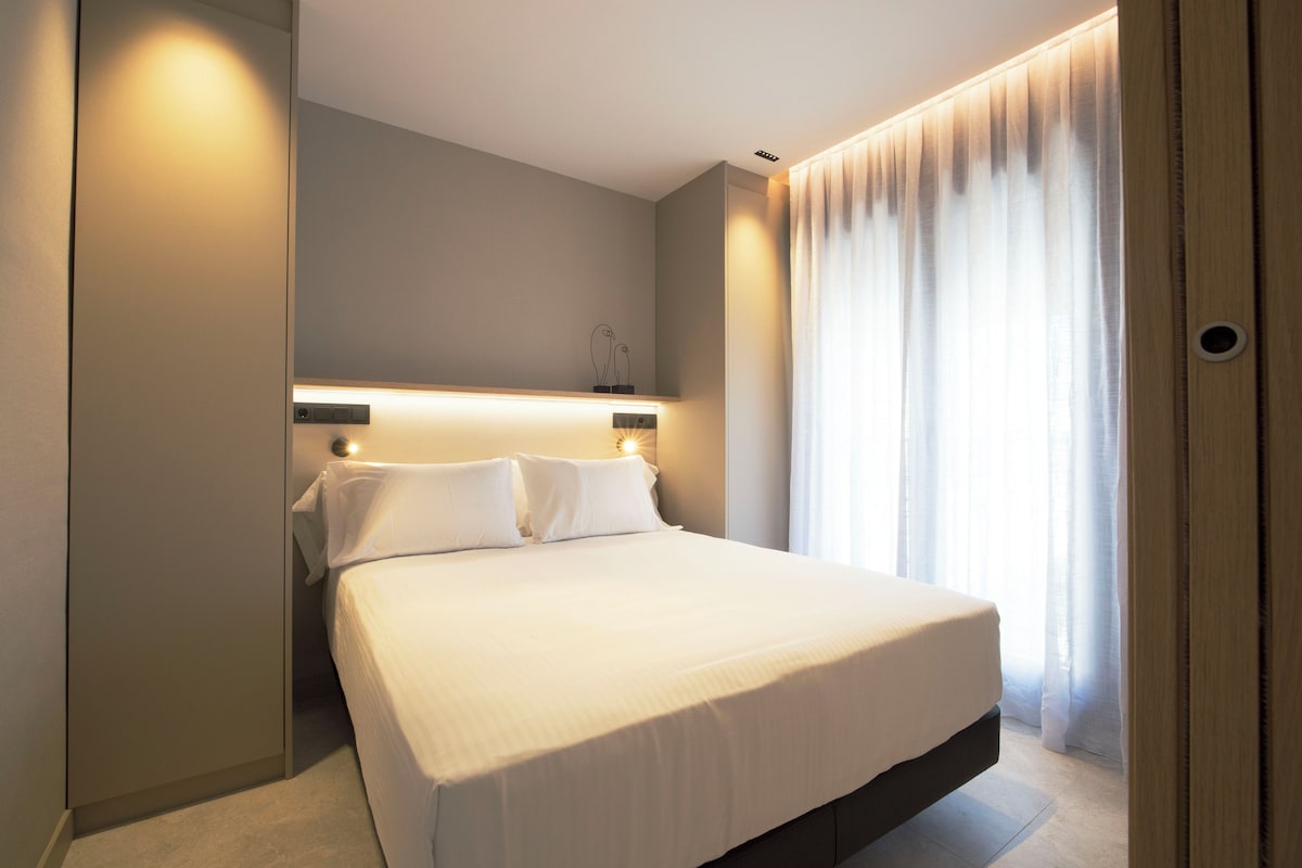 Una habitación de hotel con cama, mesita de noche, lámpara de pared, ventana con cortinas y puerta.